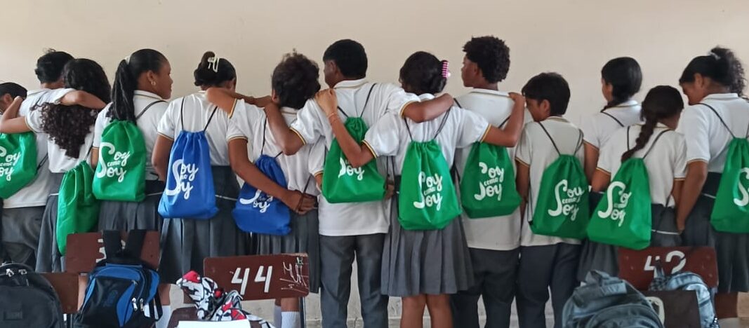 Ninos en una clase muestran sus bolsitas con la imagen de la intervencion Soy Como Soy. Han completado la intervencion y aumentada su estima corporal.

Children gather in a classroom, showing their backpacks with the Soy Como Soy logo